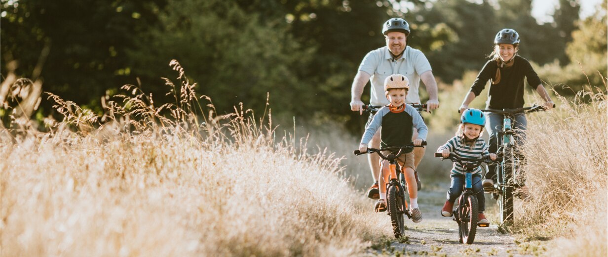 Parents and their two small children ride bicycles on a path through a field of tall grass.