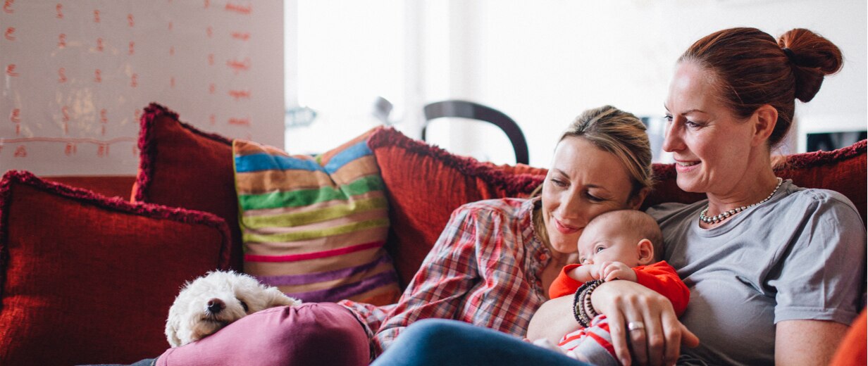 Two mothers sit on a couch, holding their baby and smiling.