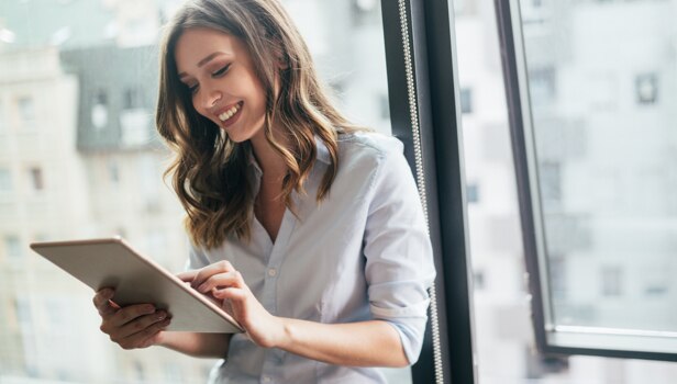A woman in a button-down shirt stands next to a well-lit office window, smiling and holding a tablet.
