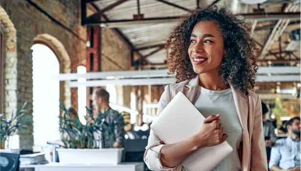 A woman who works at a tech company holds a laptop and stares into the distance, smiling.